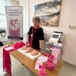 Grażyna Chaberko w czarnej sukience i ciemnoróżowej apaszce stoi przy stoisku Lubelskiego Stowarzyszenia Amazonek. Na stoliku przed nią leżą fantomy kobiecych piersi i ulotki informacyjne.
