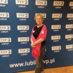 Grażyna Chaberko w ciemnych jeansach i czarnej koszulce z ciemnoróżową chustą z frędzlami pozuje na tle granatowej ścianki z logotypami TVP3 Lublin.
