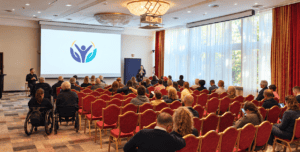 Uczestnicy siedzą na czerwonych krzesłach w sali konferencyjnej i słuchają prezentacji prowadzonej przed dużym ekranem. Wśród publiczności znajdują się również osoby korzystające z wózków. Na ekranie wyświetlono symbol przedstawiający stylizowane postacie, kojarzony z tematyką dostępności i włączenia społecznego.