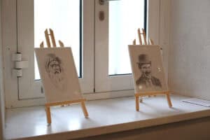 Na białym parapecie stoją dwa małe rysunki w ramach. Po lewej znajduje się portret brodatego mężczyzny w turbanie, a po prawej portret uśmiechniętego Charliego Chaplina w meloniku.