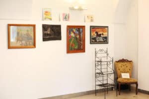 Na jasnej ścianie galerii wiszą kolorowe obrazy w drewnianych ramach oraz drewniana płaskorzeźba ze zwierzętami. W galerii stoi metalowy regał oraz antyczny fotel z tapicerką w kolorze starego złota.