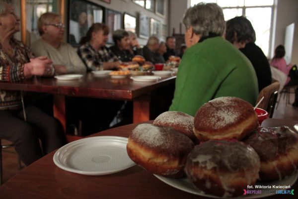 Na pierwszym planie talerz z apetycznymi pączkami, w tle Członkowie Stowarzyszenia siedzący przy stole