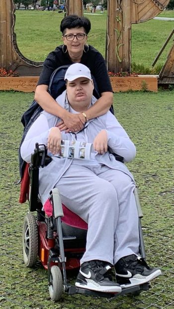 Mateusz w szarym dresie i kaszkietówce. Za nim stoi mama - szczupła, wysoka, usmiechnieta kobieta z krótkimi czarnymi włosami i w okularach , ubrana w czarną bluzę.