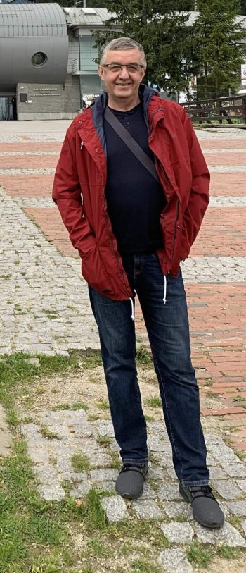 Jacek Bobruś uśmiechnięty mężczyzna w okularach, ubrany w czarną koszulkę, granatowe dżinsy i czerwoną kurtkę. stoi na tle drzew iglastych i dużego budynku.