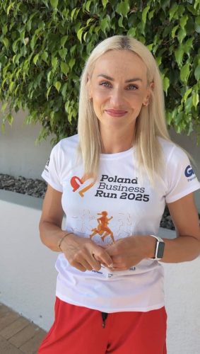 Portret uśmiechniętej lekkoatletki Justyny Święty-Ersetic w białej koszulce Poland Business Run 2025 na tle zielonych liści.
