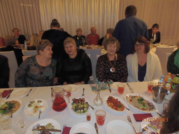 Cztery kobiety siedzące przy zastawionym stole, w tle inne osoby obu płci