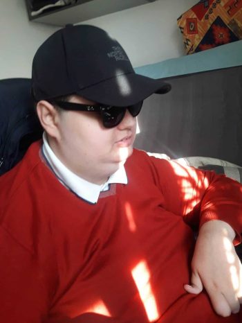 Mateusz Bobruś - młody mężczyzna - ujęcie z półprofilu - w ciemnych okularach, czarnej czapce z daszkiem, ubrany w czerwoną bluzę i koszulkę z białym kołnierzykiem. Na jego twarzy odbijają się promienie słońca.